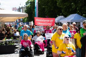 Bilde fra Stolthetsparaden 2019, mange mennesker som går eller sitter i rullestol. Noen bærer faner med kamprop om likestilling. De fleste på bildet har på seg t-skjorter i gult, blått eller rosa hvor det står "Stolt, sterk synlig"