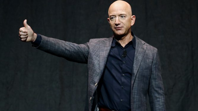Bildet viser Amazon-grunnlegger Jeff Bezos kledd i svart og grått mot svart bakgrunn. Han har den høyre hånden strukket ut og gir tommel opp.