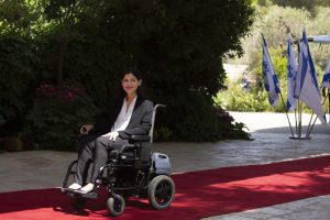 Bildet viser Karine Elharrar sittende i rullestolen sin på en rød løper. Hun smiler. I bakgrunnen står det israelske flagg.