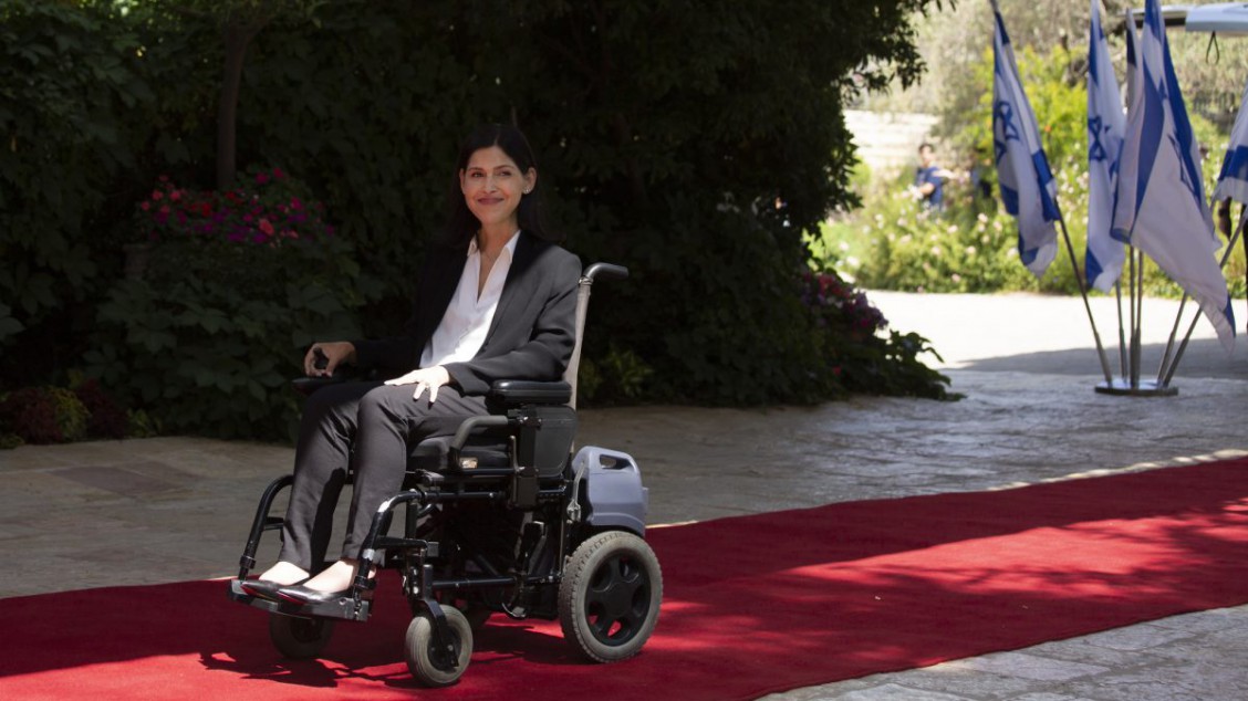 Bildet viser Karine Elharrar sittende i rullestolen sin på en rød løper. Hun smiler. I bakgrunnen står det israelske flagg.