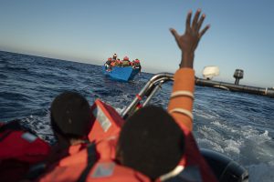 Bildet viser i bakgrunnen en blå båt fylt av mennesker. I forgrunnen er bildet uklart, viser to mennesker med redningsvest. Den ene strekker hånden i været.