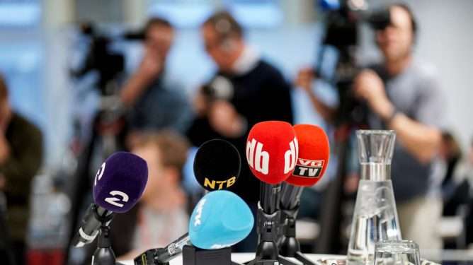 Pressekonferanse: Mikrofoner fra flere medier plassert på et bord. I bakgrunnen (uklart) tre mennesker med kamera og mikrofon.