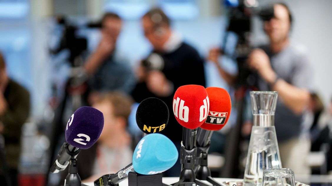 Pressekonferanse: Mikrofoner fra flere medier plassert på et bord. I bakgrunnen (uklart) tre mennesker med kamera og mikrofon.