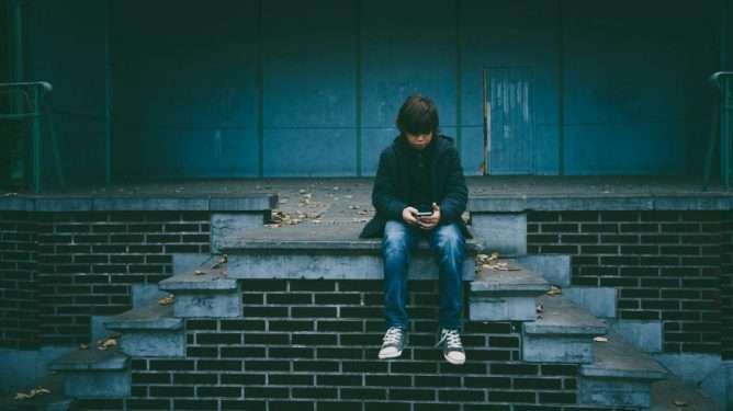 En gutt sitter alene i en trapp med en mobiltelefon i hånda.