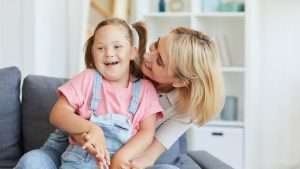 En jente med downs syndrom sitter på fanget til moren sin. Jenta ler og moren gir henne en klem.