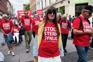 Bilde fra stolthetsparaden, viser mennesker som går i tog, alle har på røde t-skjorter der det står "stolt, sterk, synlig".
