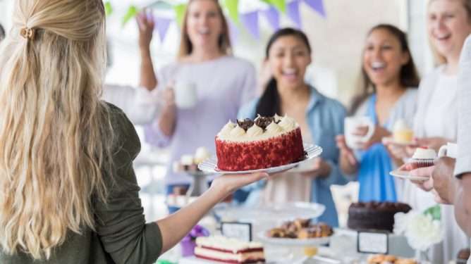 En kvinne i fokus med ryggen til som viser frem en kake. I bakgrunnen, ute av fokus: fire kvinner som ser imponert ut over kaken.